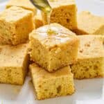 Honey being spooned on top of squares of cornbread stacked on a white platter