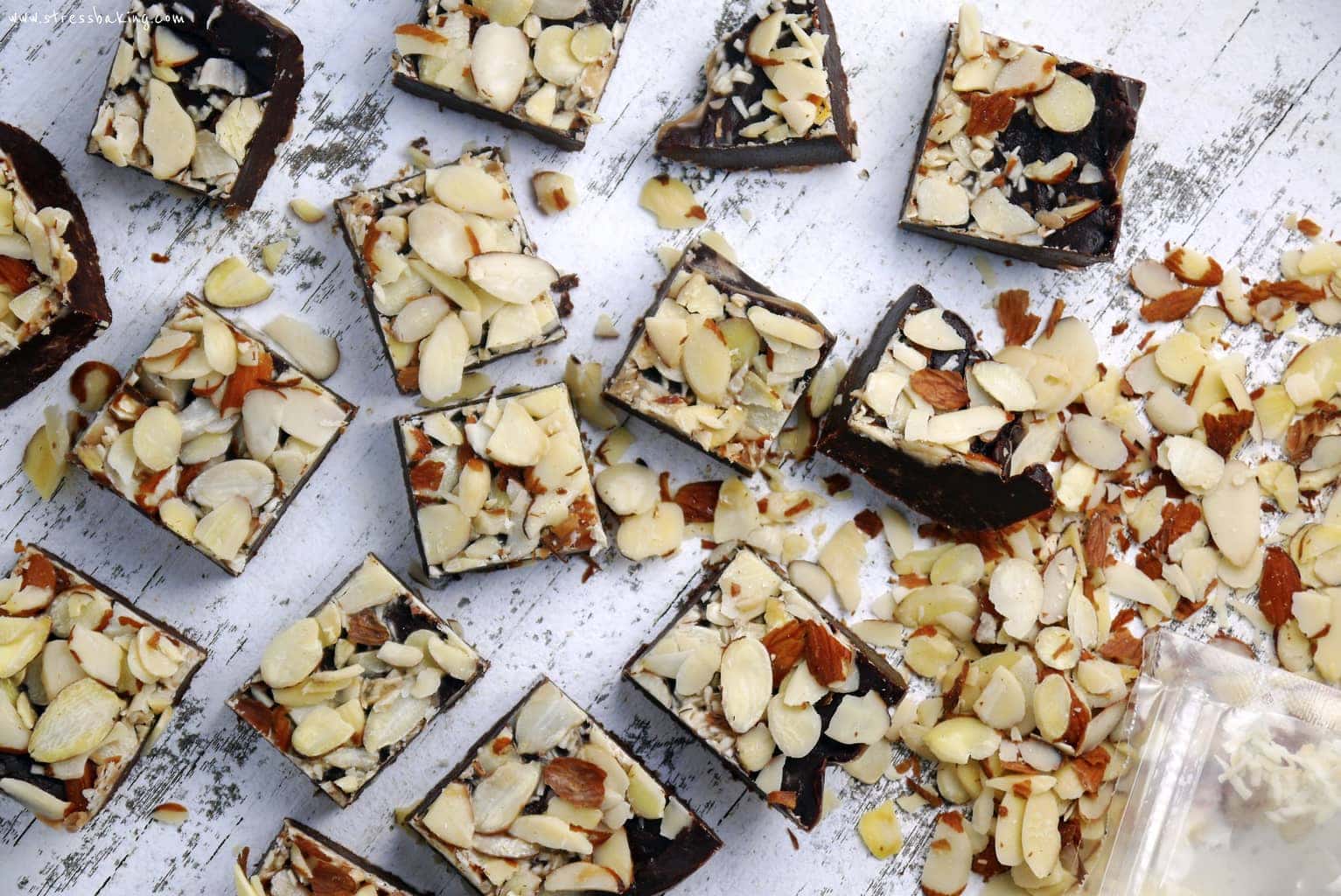 Squares of chocolate fudge topped with sliced almonds strewn about a distressed white surface