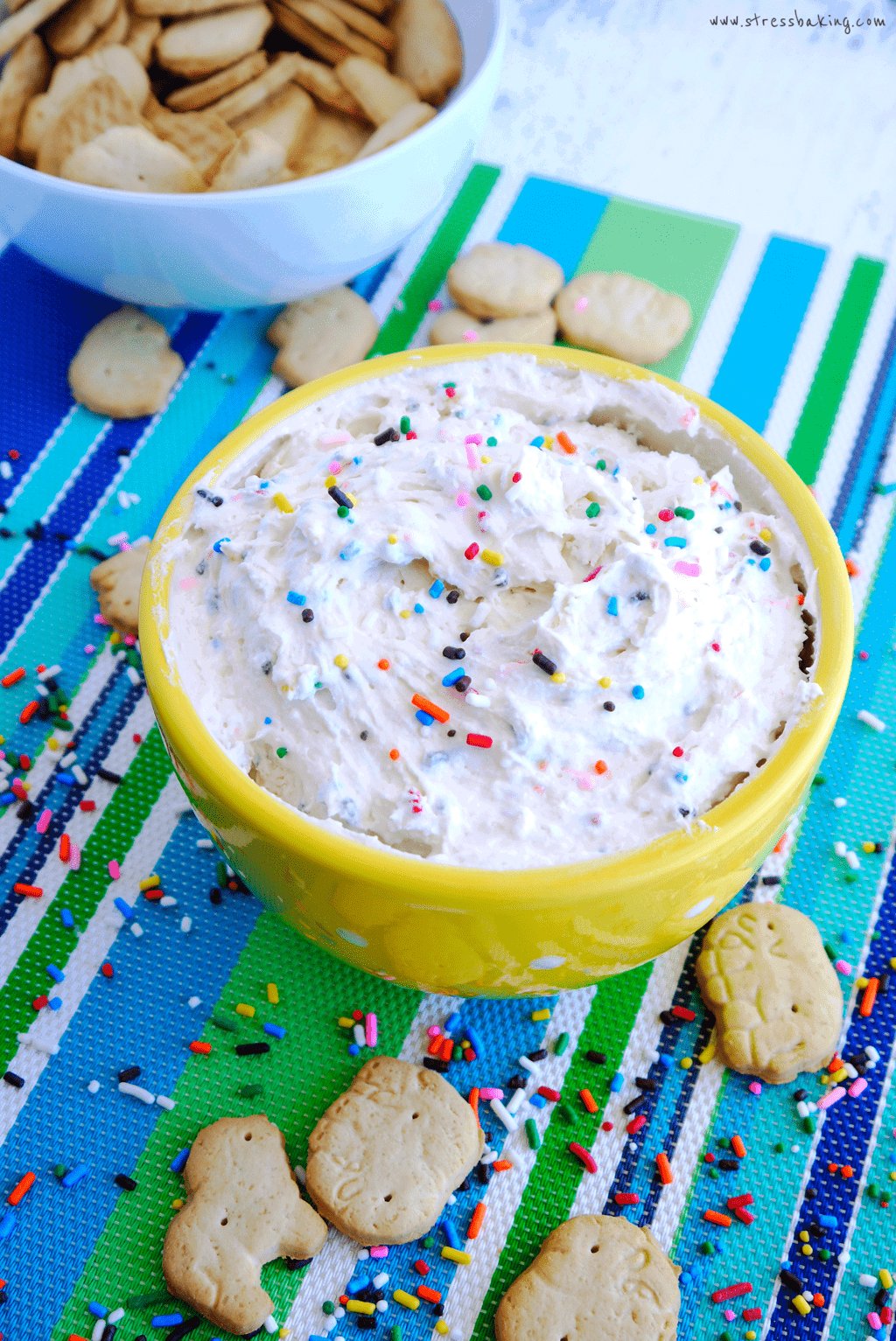 Yellow bowl full of funfetti dunkaroo dip topped with sprinkles on a colorful striped placemat with animal crackers