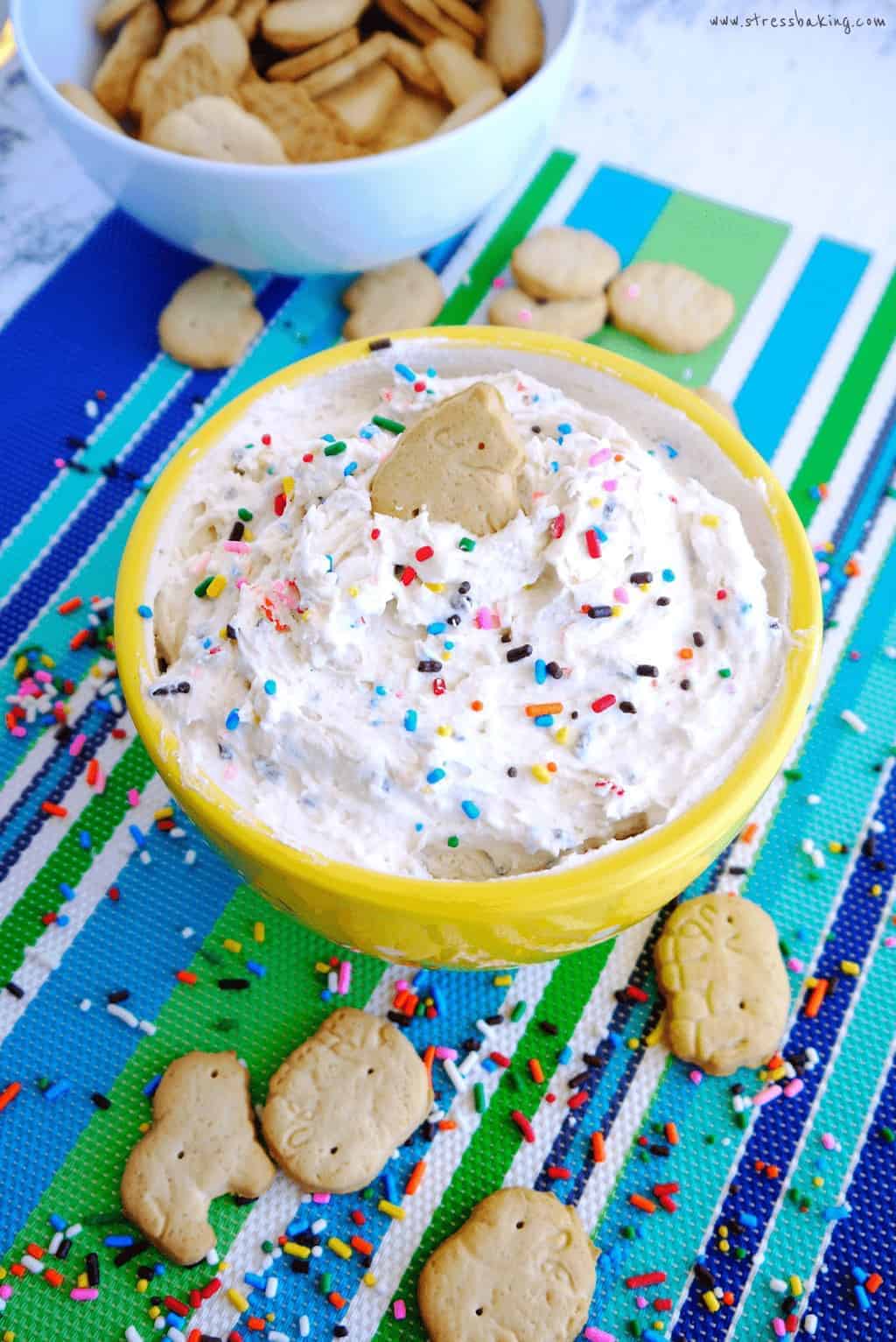 An animal cracker dipped in a yellow bowl full of funfetti dunkaroo dip topped with sprinkles