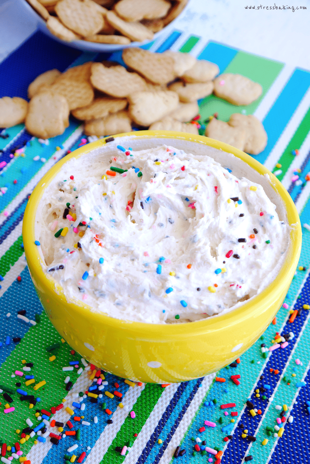 Yellow bowl full of funfetti dunkaroo dip topped with sprinkles on a colorful placemat with animal crackers