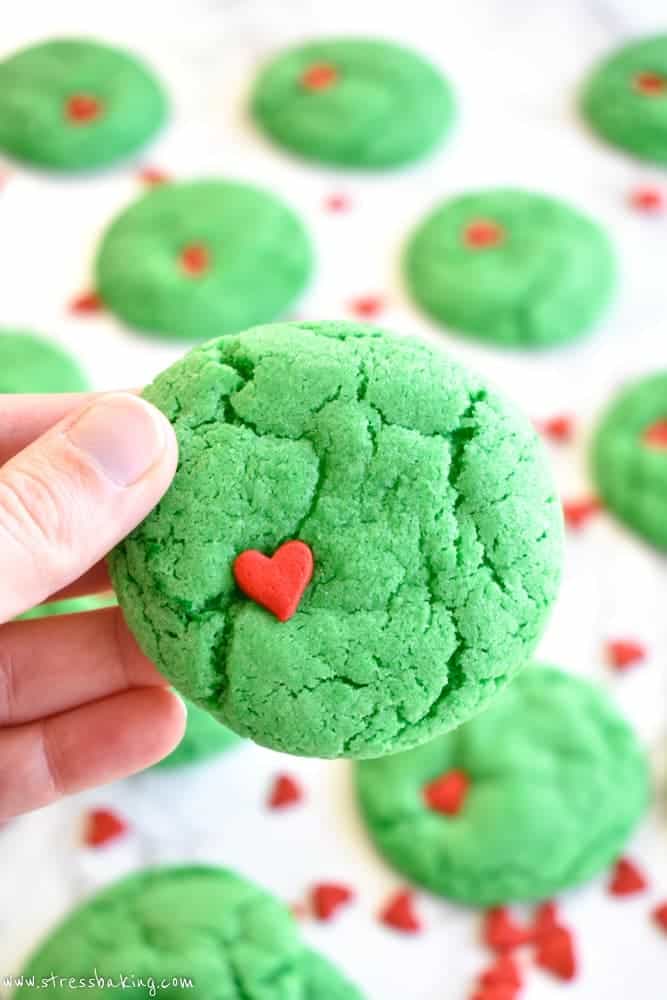 Hand holding a vibrant green Grinch cookie with a red heart sprinkle held in front of other cookies