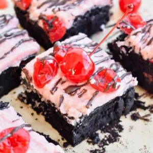Brownies topped with pink frosting and maraschino cherries and chocolate drizzle