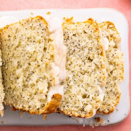 A glazed lemon poppy seed loaf with pieces falling to the side to reveal the moist yellow inside dotted with black poppy seeds on a pink surface