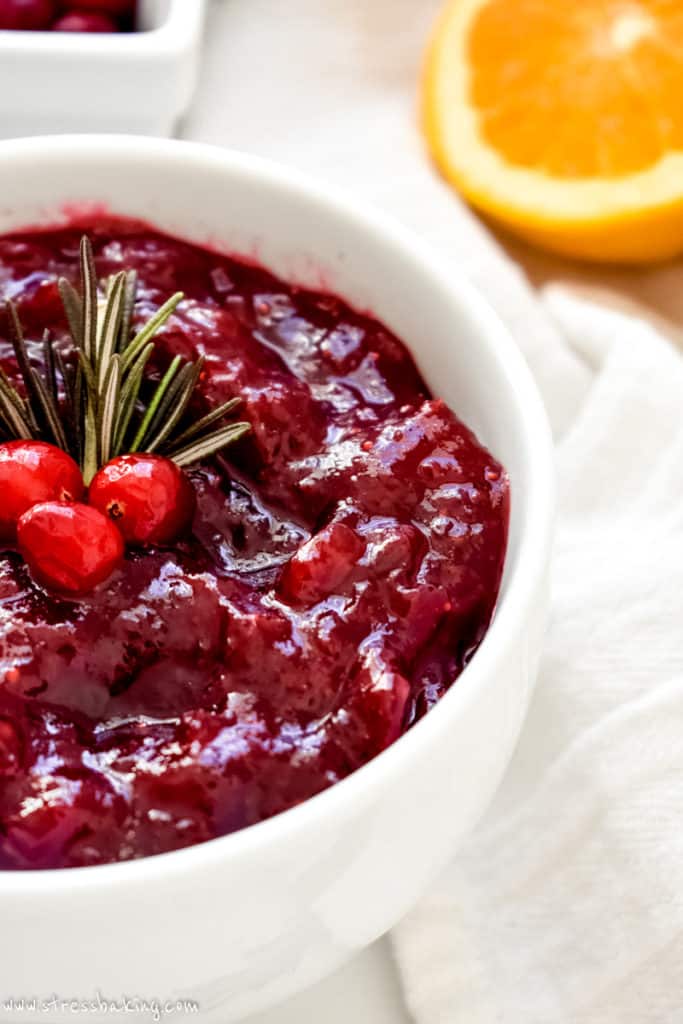 Close up of brightly colored cranberry sauce in a white bowl topped with cranberries and rosemary sprigs