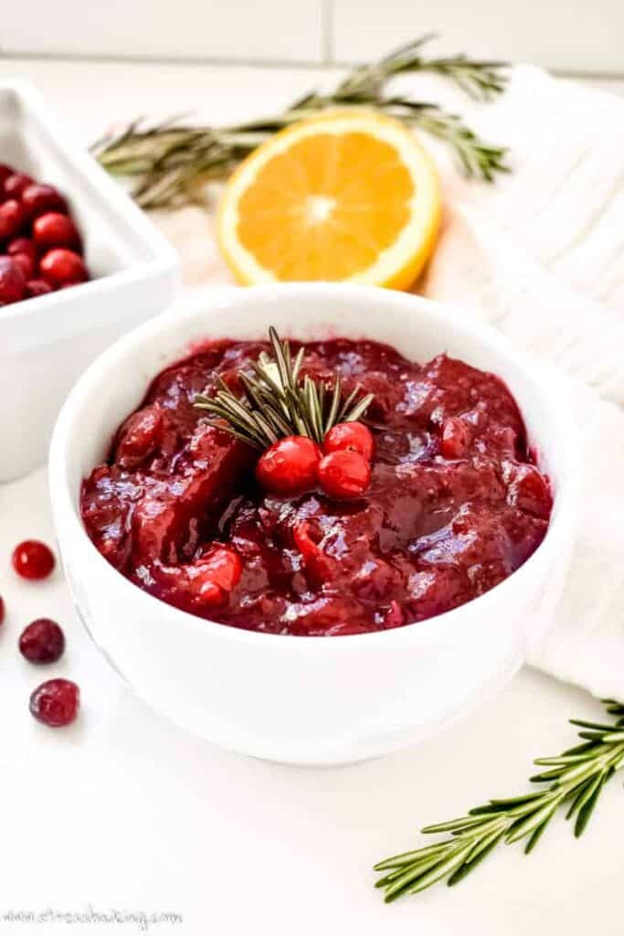 Brightly colored cranberry sauce in a white bowl topped with cranberries and rosemary sprigs in front of half an orange