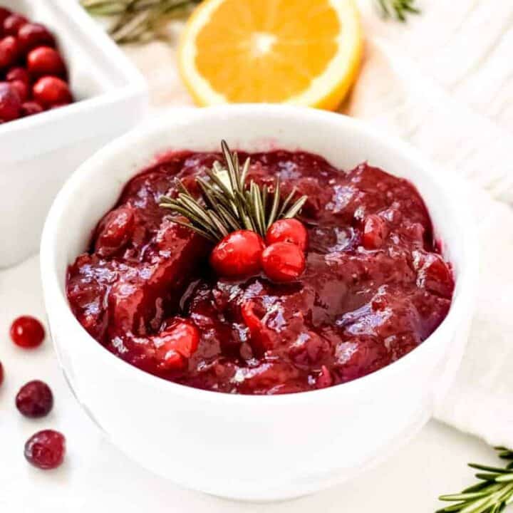 Brightly colored cranberry sauce in a white bowl topped with cranberries and rosemary sprigs in front of half an orange