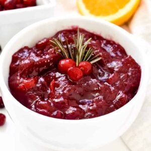 Brightly colored cranberry sauce in a white bowl topped with cranberries and rosemary sprigs