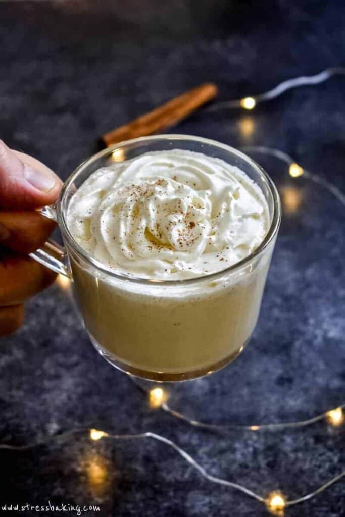 A hand holding a clear mug filled with creamy homemade eggnog and topped with whipped cream and nutmeg