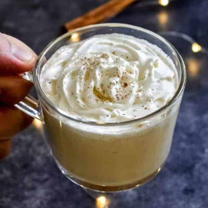 A hand holding a clear mug filled with creamy homemade eggnog and topped with whipped cream and nutmeg