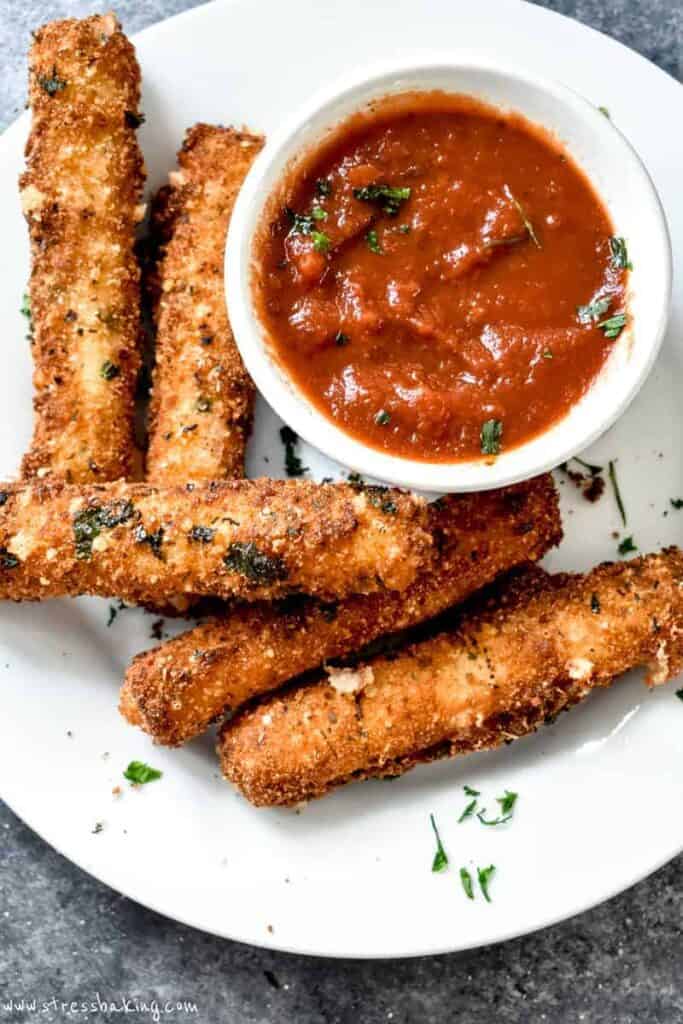 Overhead shot of browned mozzarella sticks on a white plate with a dish of marinara sauce