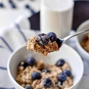 A spoonful of blueberry overnight oats held over a white bowl next to a glass of milk