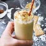 A hand holding a coconut cream pie milkshake topped with toasted coconut and graham cracker crumbles