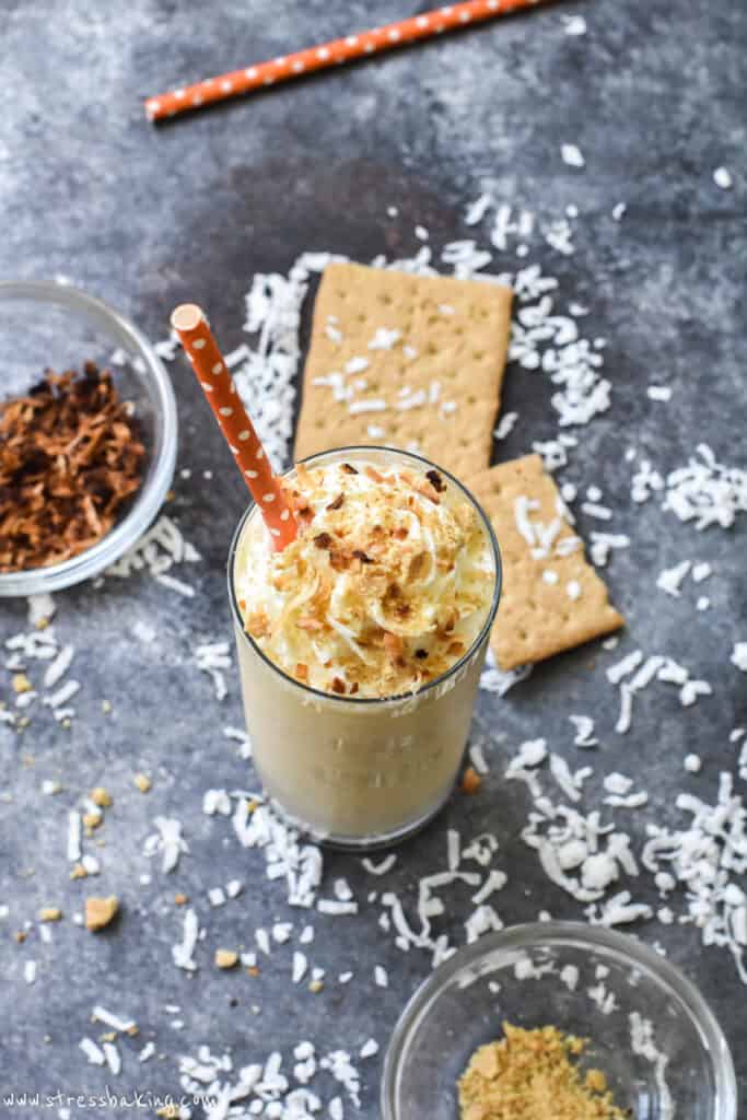 A coconut cream pie milkshake topped with toasted coconut and graham cracker crumbles with an orange straw