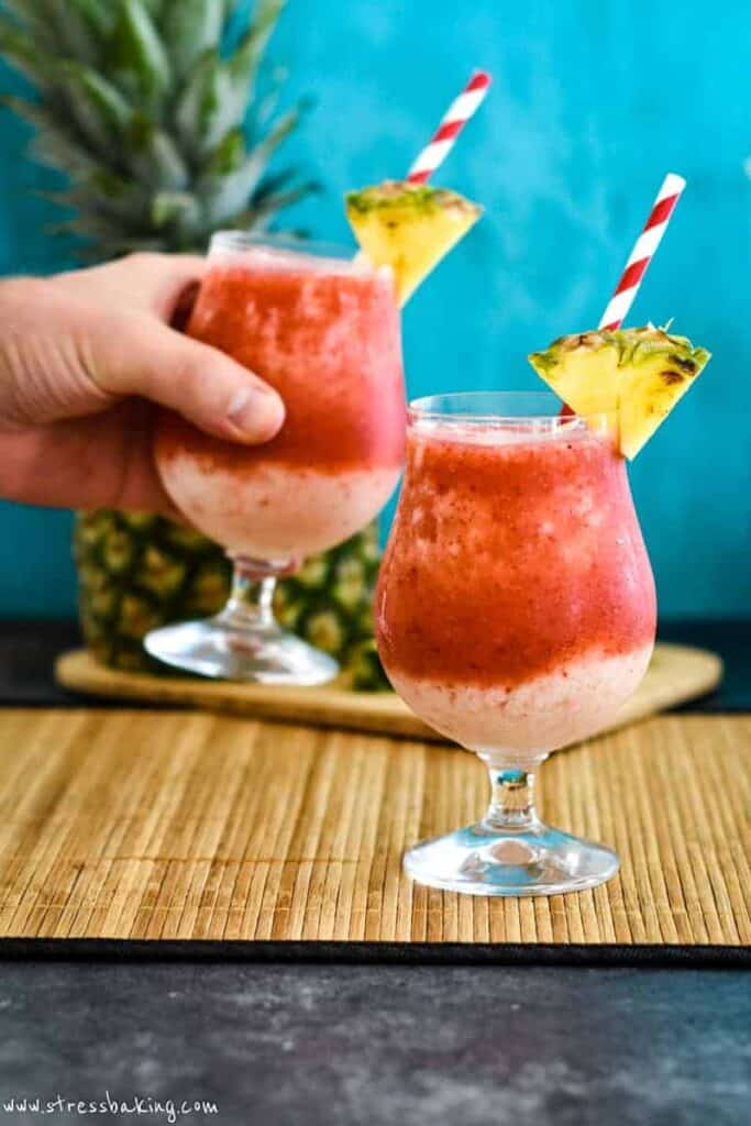 Two vibrant red and cream colored frozen drinks with wedges of pineapple with one being lifted