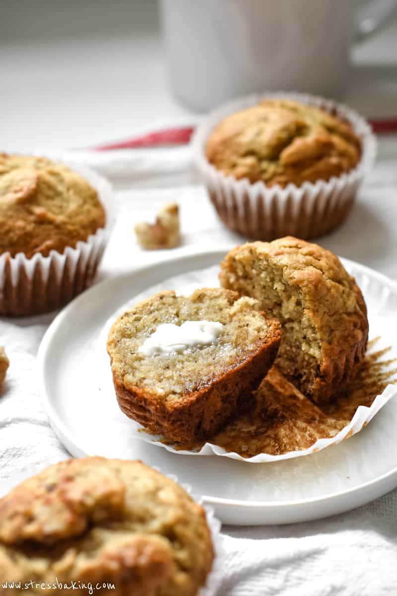 This photo shows a banana muffin on a plate cut in half with butter. 3 muffins are around the plate on a kitchen counter.