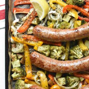 Overshot photo of browned sausages on top of a sheet pan of seasoned roasted veggies