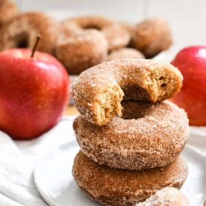 A stack of apple cider donuts on a white plate with red apples