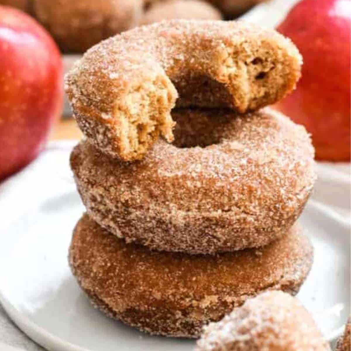 A stack of apple cider donuts on a white plate with red apples