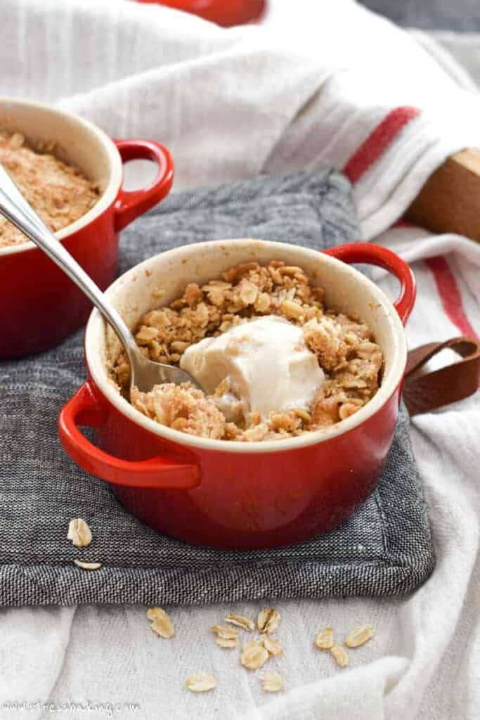 A mini cocotte full of freshly baked apple crisp and topping with a spoon digging into scoop of ice cream