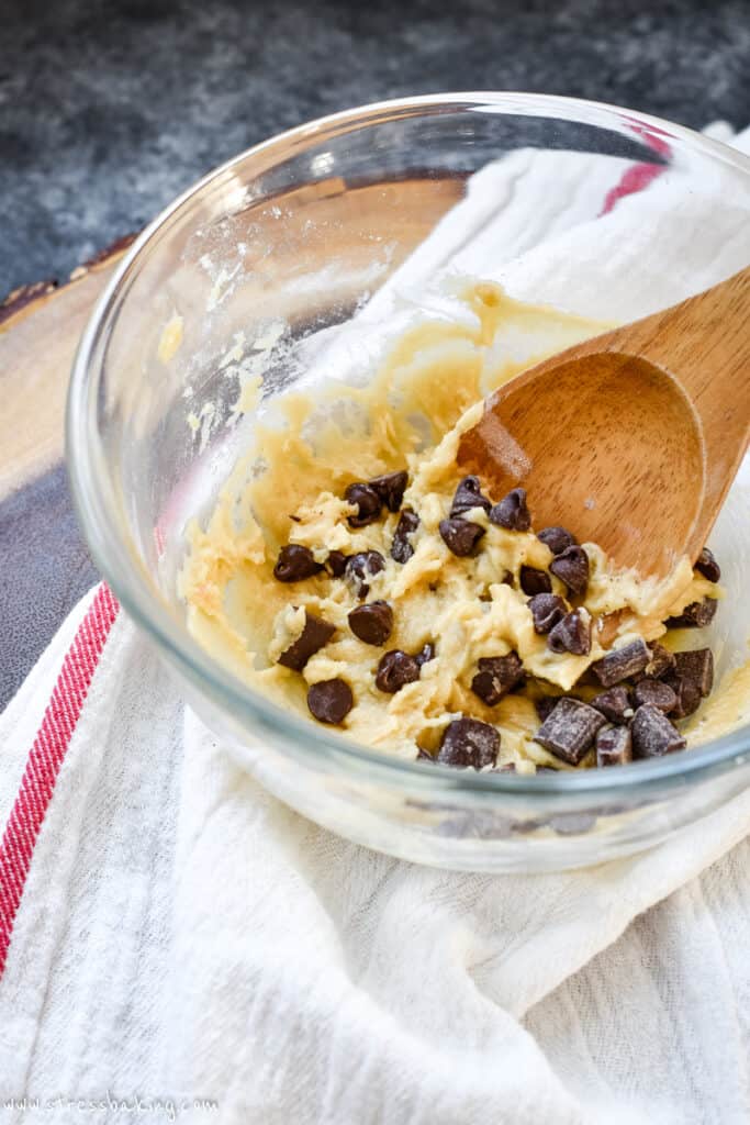 A glass bowl of buttery chocolate chip cookie dough with a wooden spoon