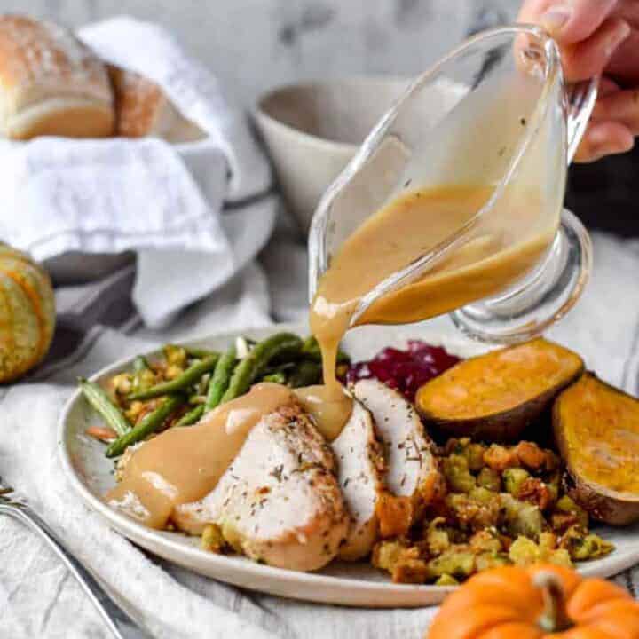Gravy being poured onto thick slices of turkey breast surrounded by Thanksgiving foods