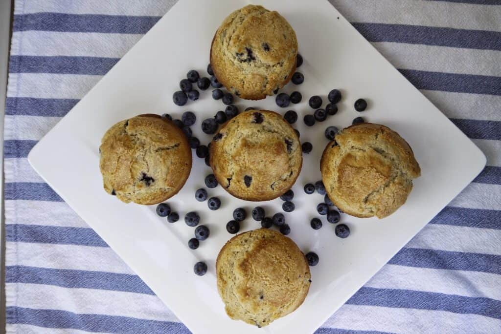 5 large blueberry muffins on a white plate with fresh blueberries