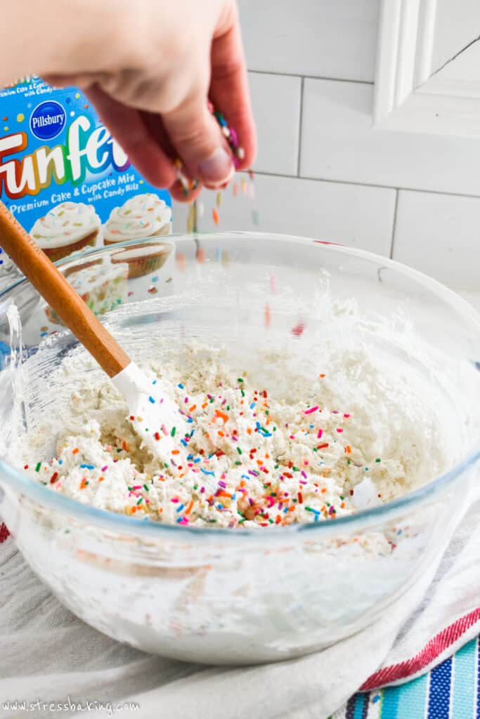 A hand dropping rainbow sprinkles into a mixing bowl