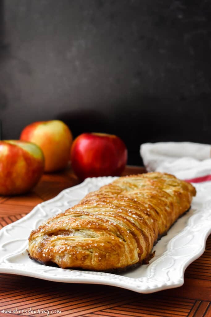 Golden brown apple strudel on a white decorative platter with apples in the background