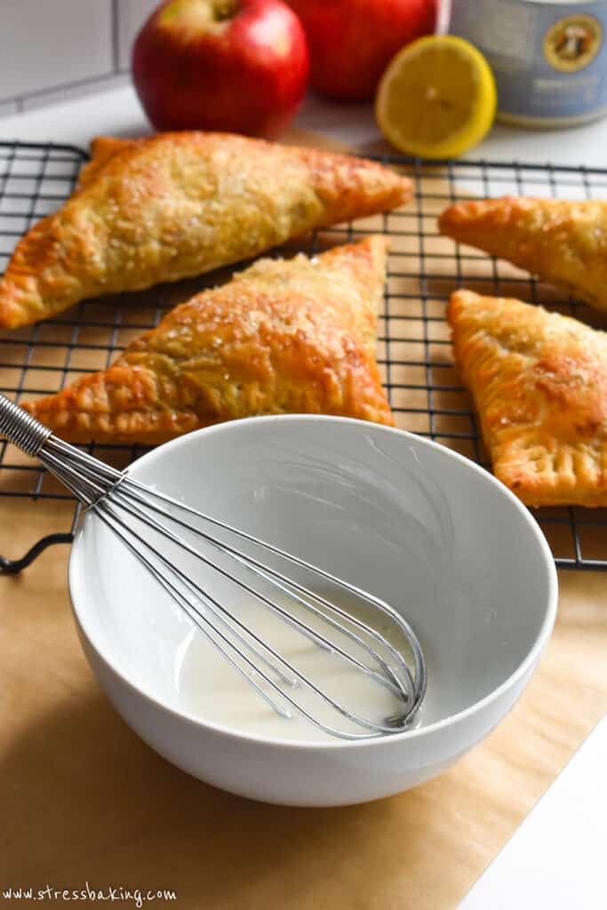 Icing in a white bowl with a small whisk in front of a wire rack of apple turnovers