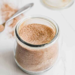A glass jar of cinnamon sugar on a white counter with a spoonful in the background