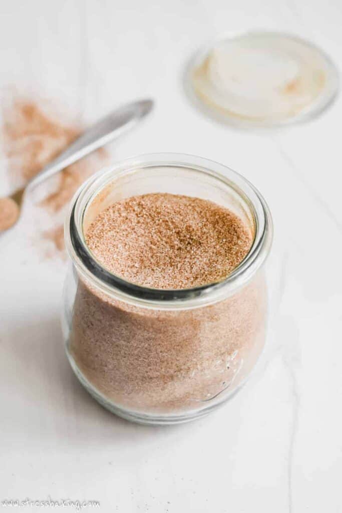 A glass jar of cinnamon sugar on a white counter with a spoonful in the background