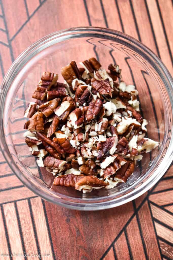 A clear bowl of toasted pecan pieces on a retro wooden surface