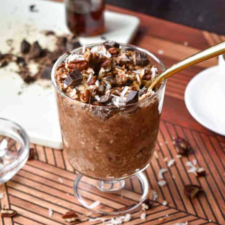 A small glass trifle dish with chocolate oats topped with pecans, coconut and chocolate chunks next to a cup of espresso
