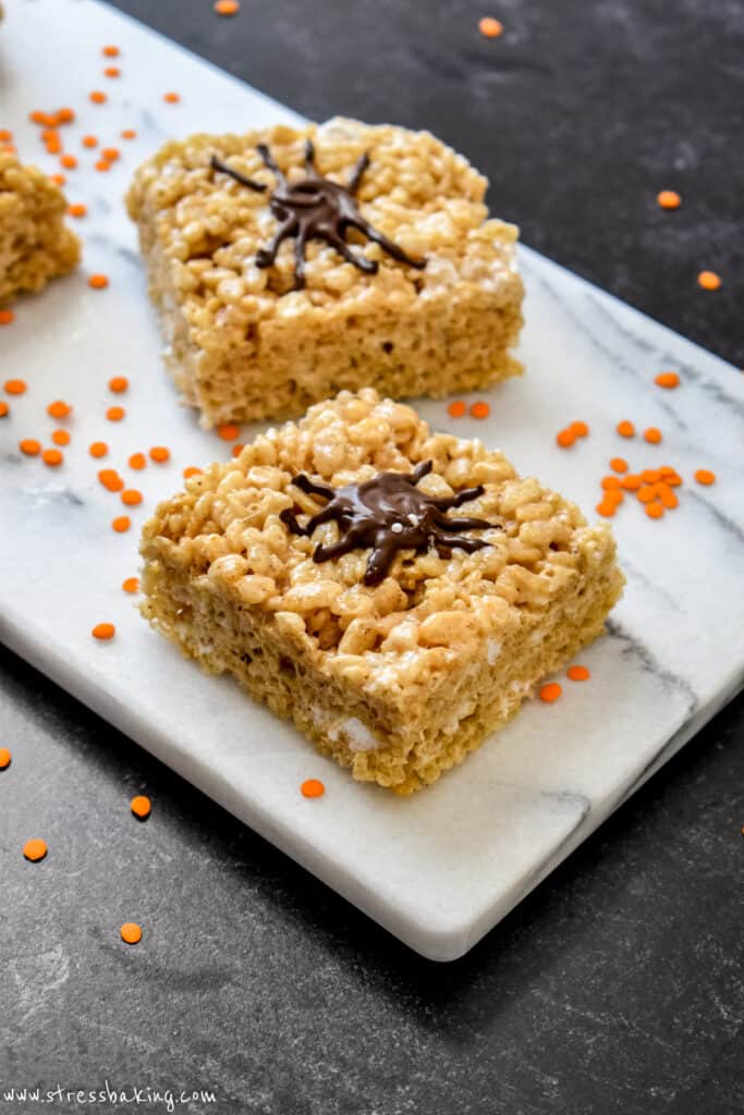 Halloween rice krispie treats with chocolate spiders decorating the tops surrounded by orange sprinkles