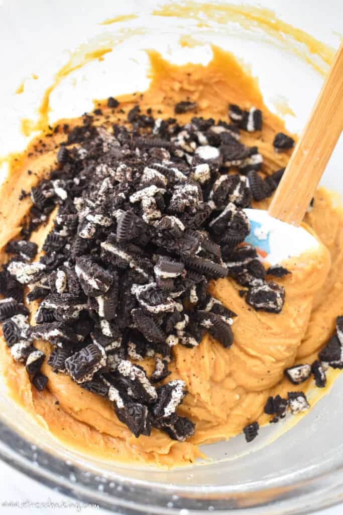 Orange colored fudge dough in a clear mixing bowl with crushed Oreos
