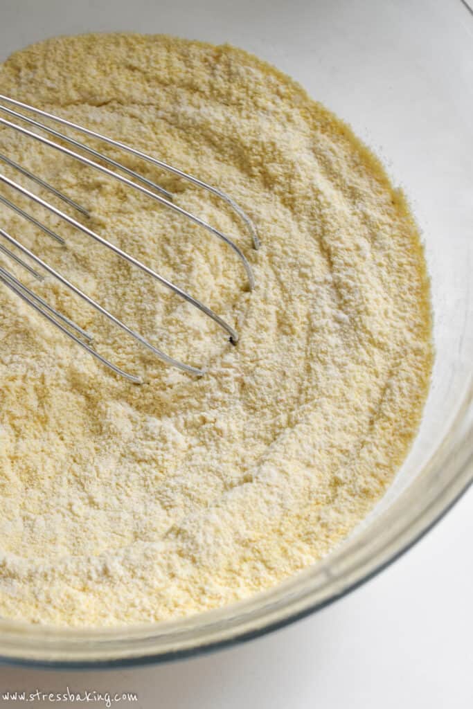 A clear mixing bowl full of flour and cornmeal and a whisk
