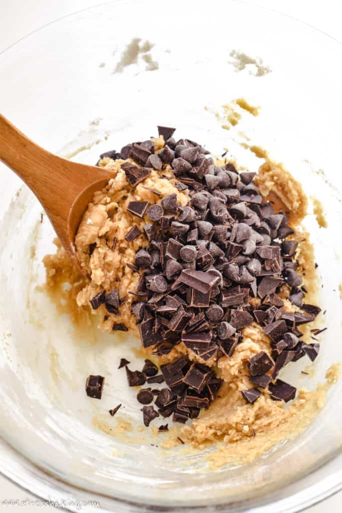 Chocolate chip cookie dough with chocolate about to be mixed in in a clear mixing bowl