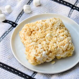 Single rice krispie treat square on a white plate atop a black and white dish towel