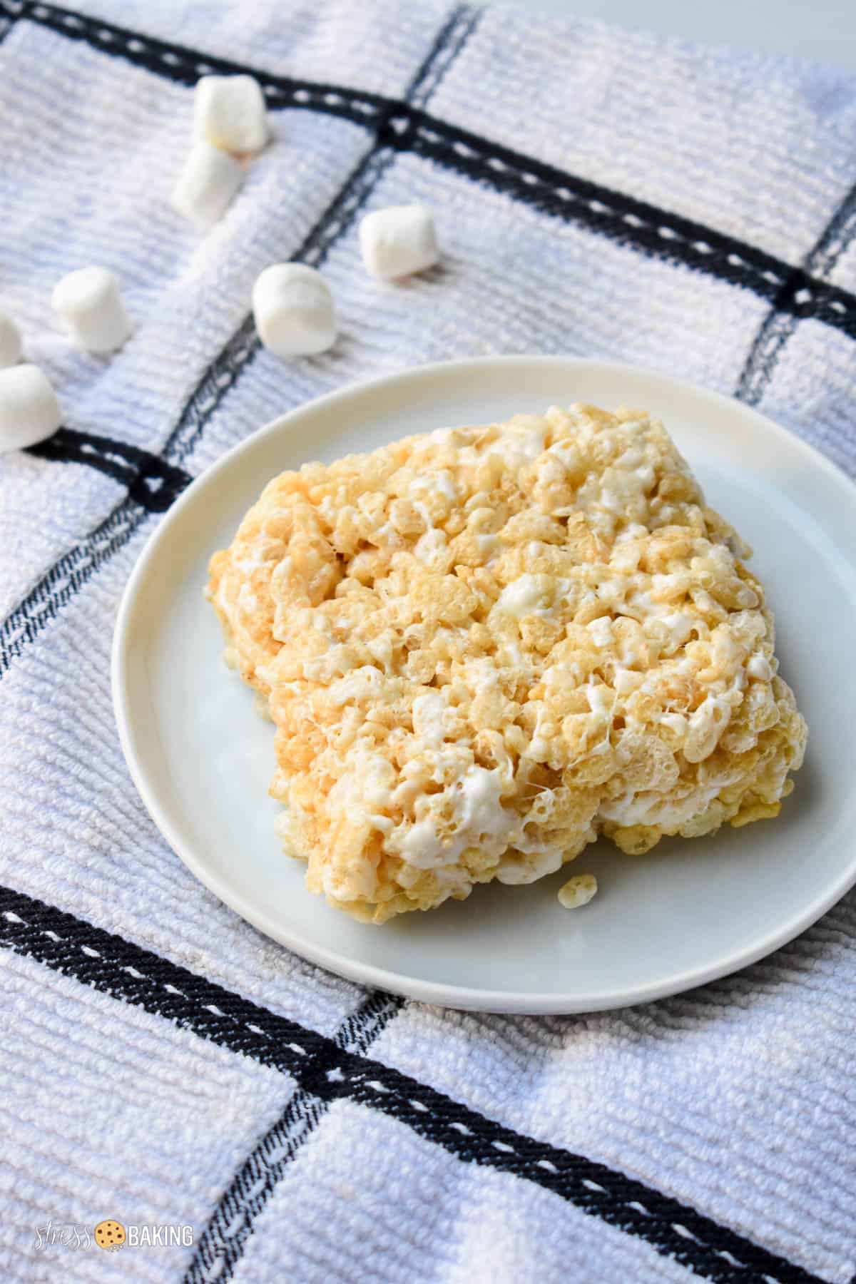 Single rice krispie treat square on a white plate atop a black and white dish towel