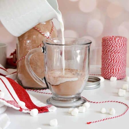 Milk being poured into a clear mug of hot cocoa mix against a blush colored background