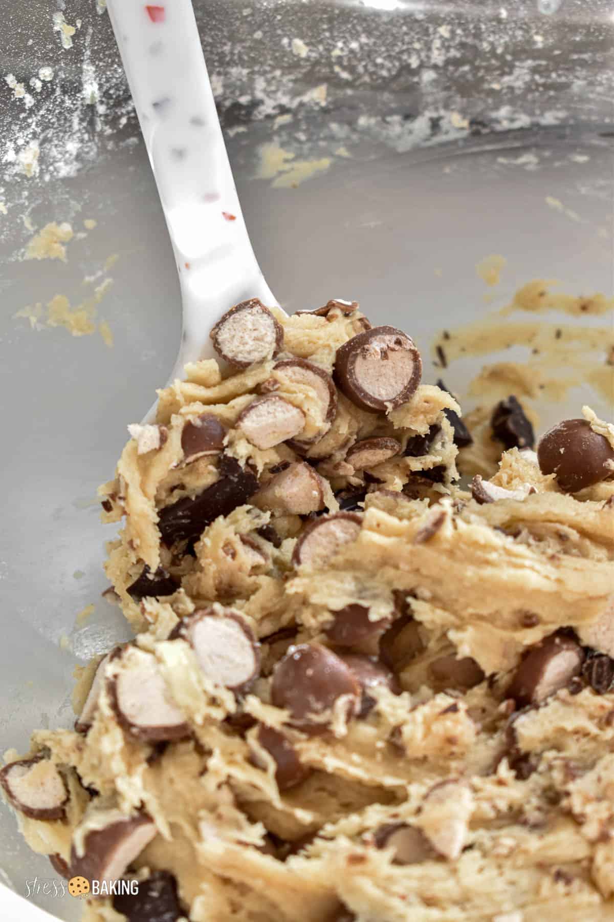 Cookie dough mixed with chopped chocolate and Whoppers candy in a clear mixing bowl