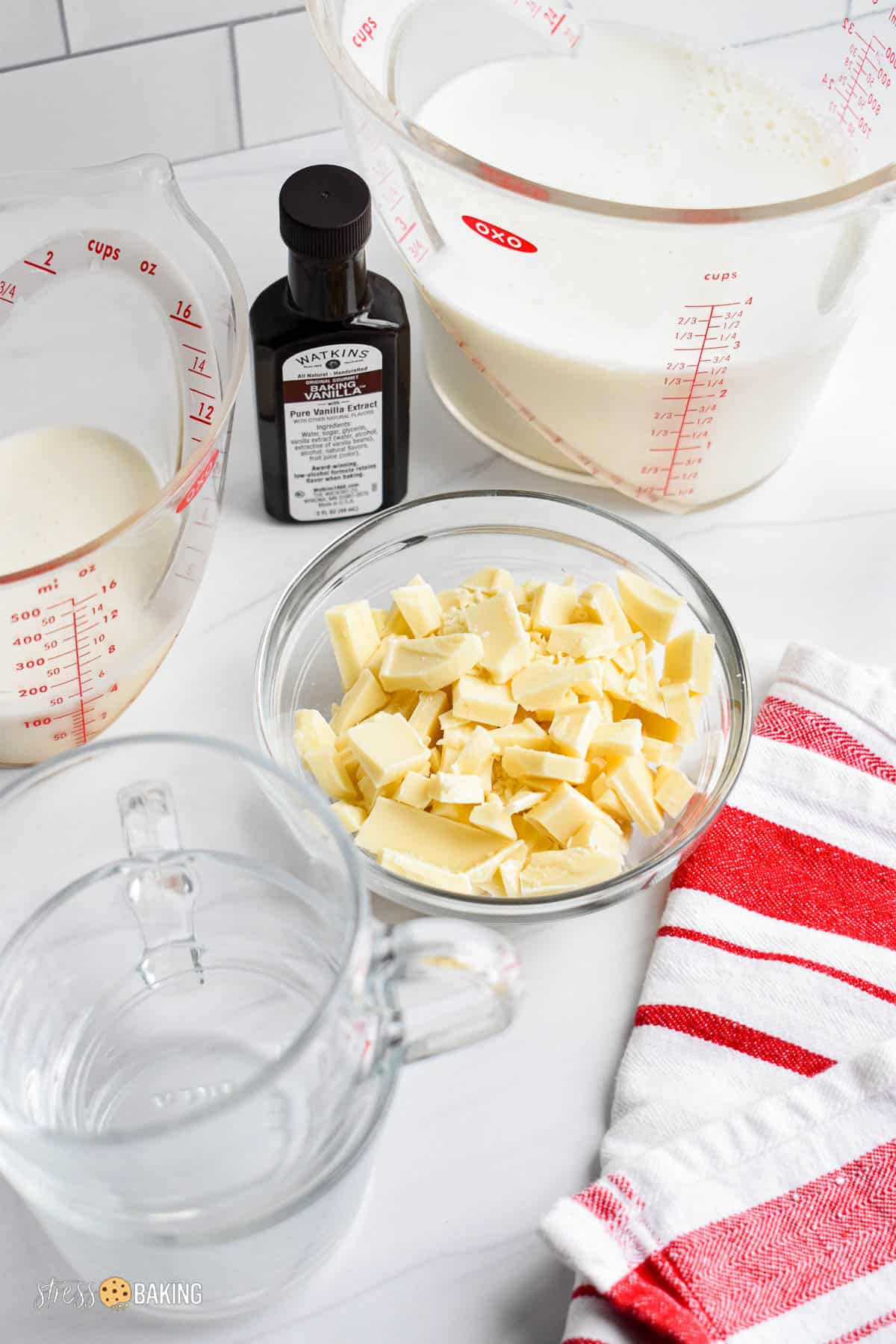 Clear bowl of chopped white chocolate with two measuring cups of milk and cream, a bottle of vanilla extract and clear mugs