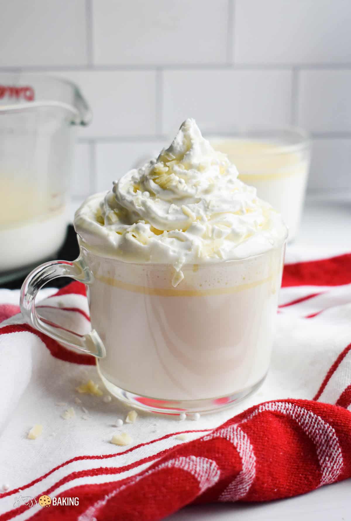 White hot chocolate topped with a mountain of whipped cream in a clear mug on a red and white dish towel