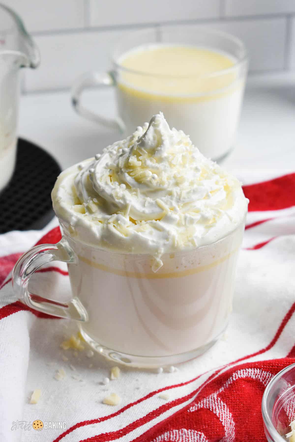 White hot chocolate topped with a mountain of whipped cream in a clear mug on a red and white dish towel