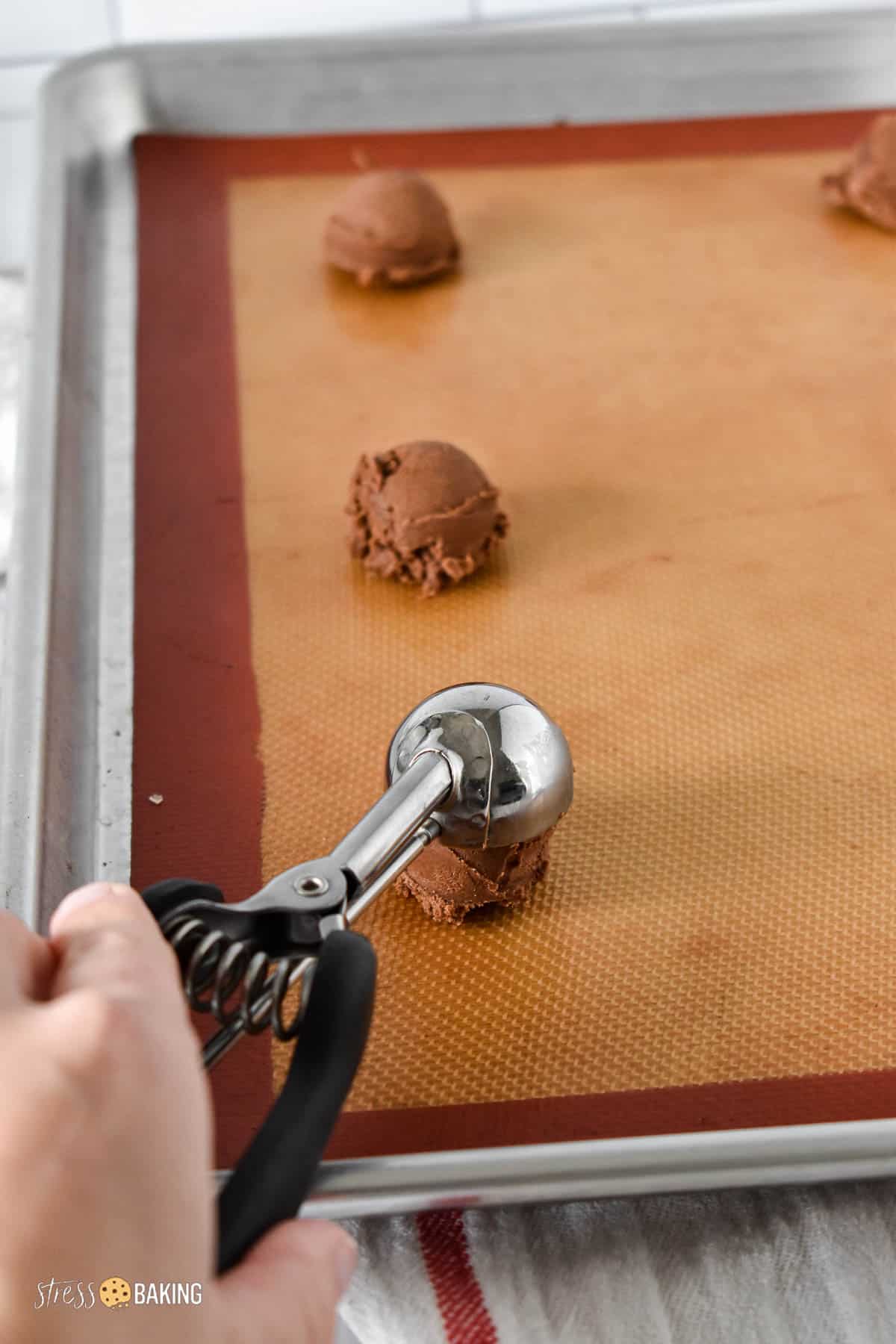 A small cookie scoop portioning out chocolate cookie dough on a baking sheet
