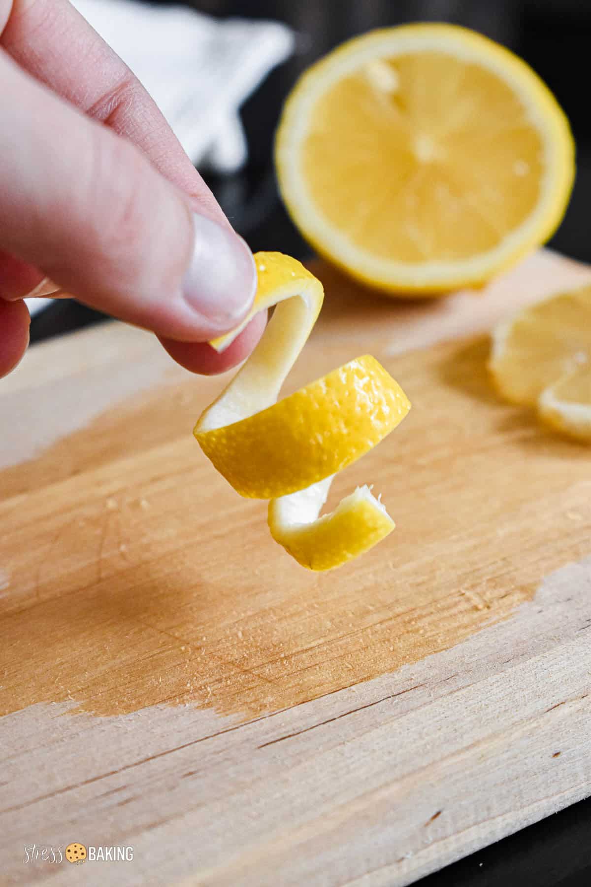 A curl of lemon twist garnish being held above a small wooden cutting board