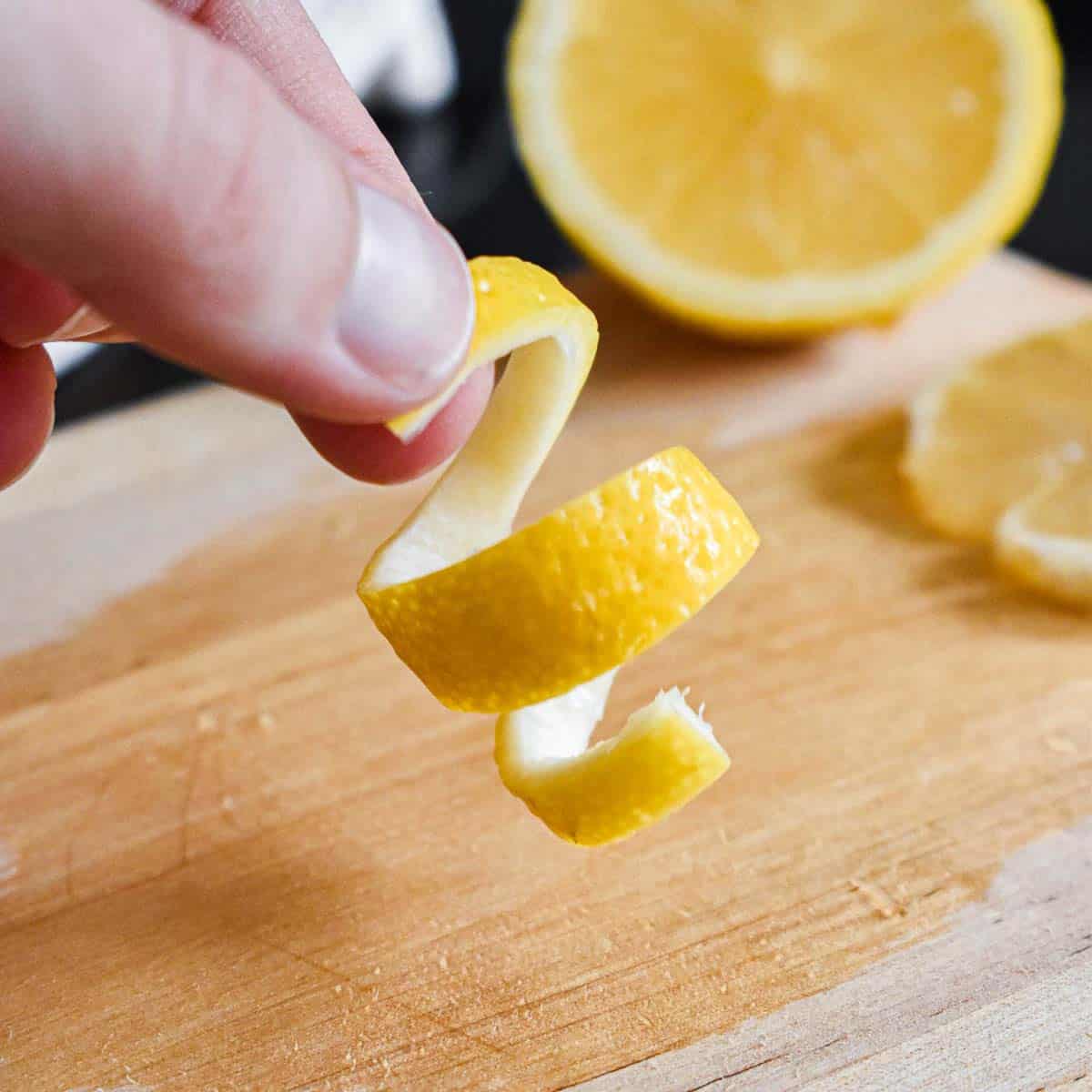 A curl of lemon twist garnish being held above a small wooden cutting board
