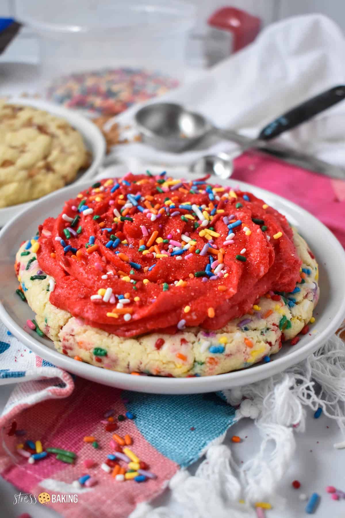 A big sugar cookie decorated with red buttercream frosting and rainbow sprinkles on a small white plate