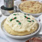 A big sugar cookie decorated with buttercream frosting and holly sprinkles on a small white plate